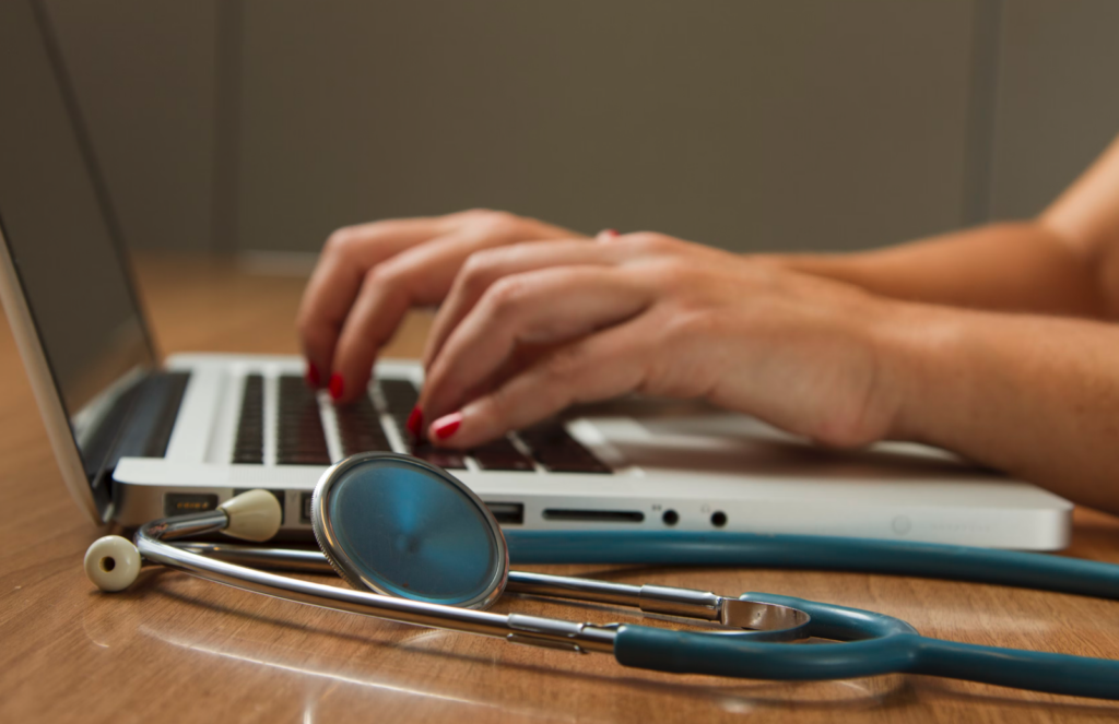 A person typing on a laptop with a stethoscope placed on the desk nearby.